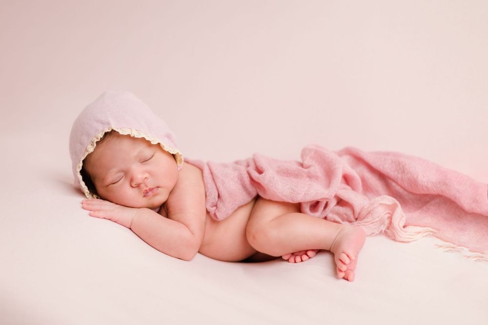 Peaceful sleeping newborn baby wearing pink bonnet draped with soft pink blanket on cream background.