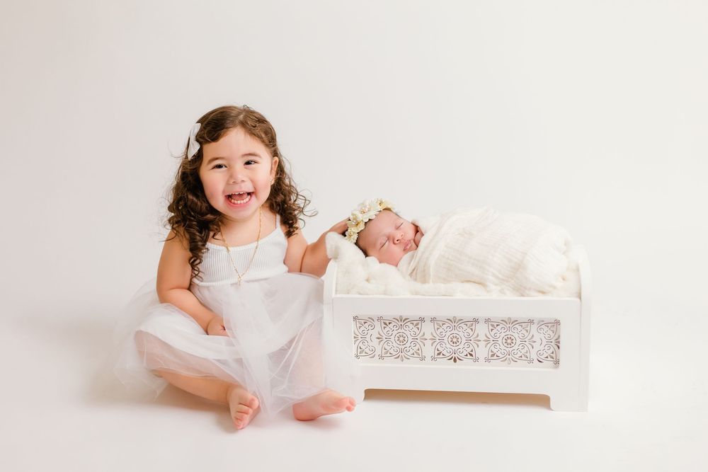 Happy toddler girl in white dress laughing beside newborn baby sleeping in white lace crib.