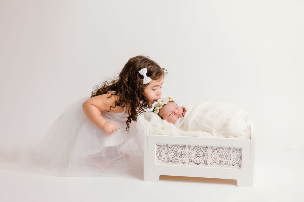 Young girl in white tutu dress leaning over to kiss sleeping newborn baby in white lace crib.