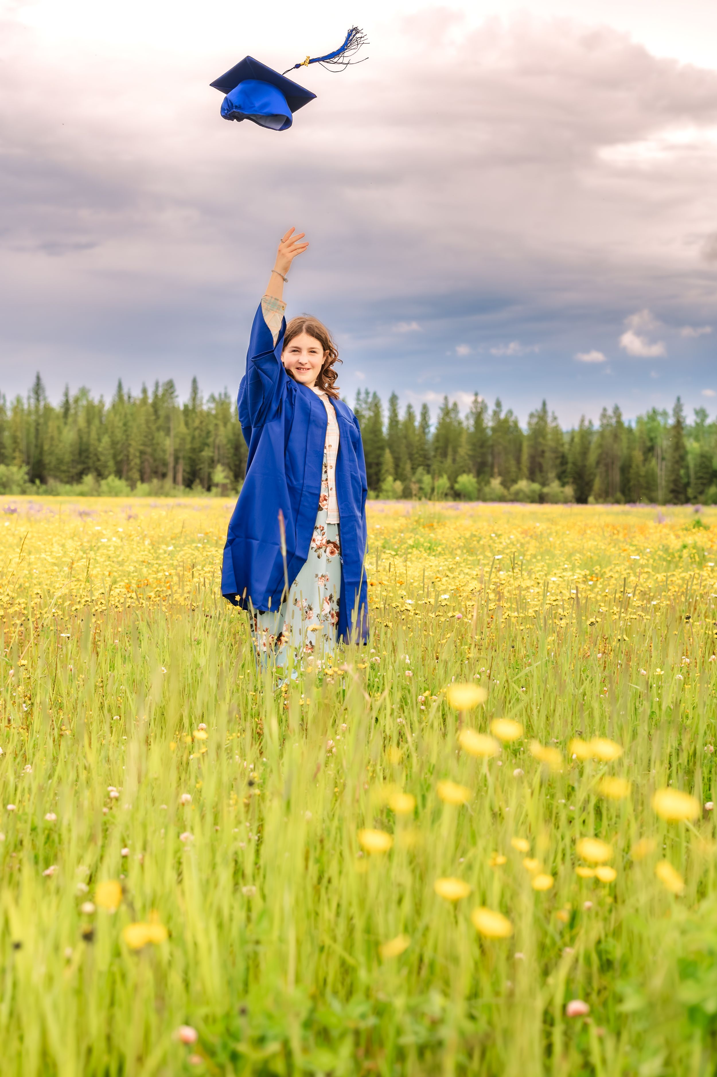 A graduate in blue cap and gown celebrates in a sunny wildflower meadow with yellow blooms and evergreen trees.