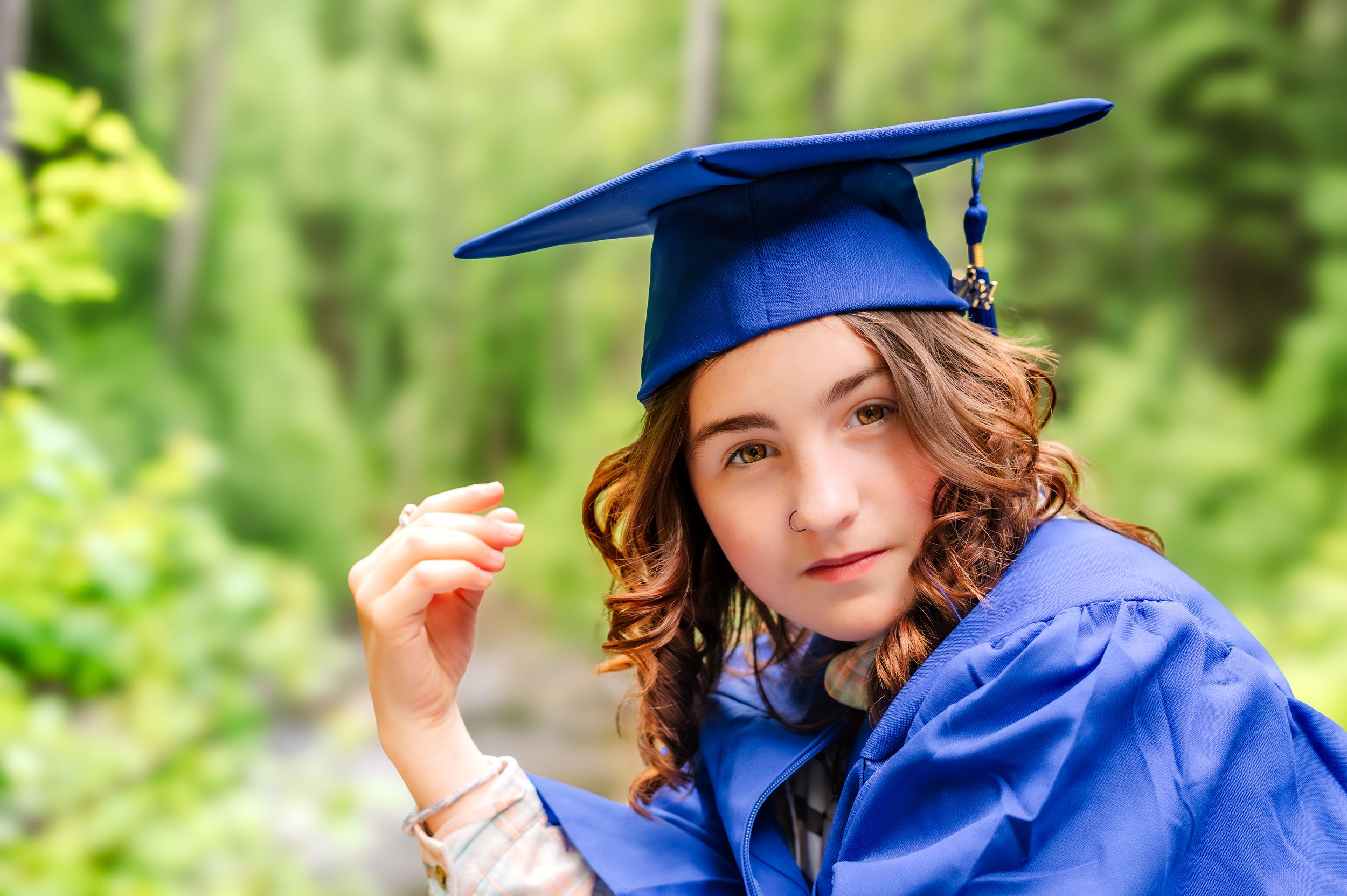 A graduate in blue cap and gown poses outdoors with a diploma while surrounded by green foliage.