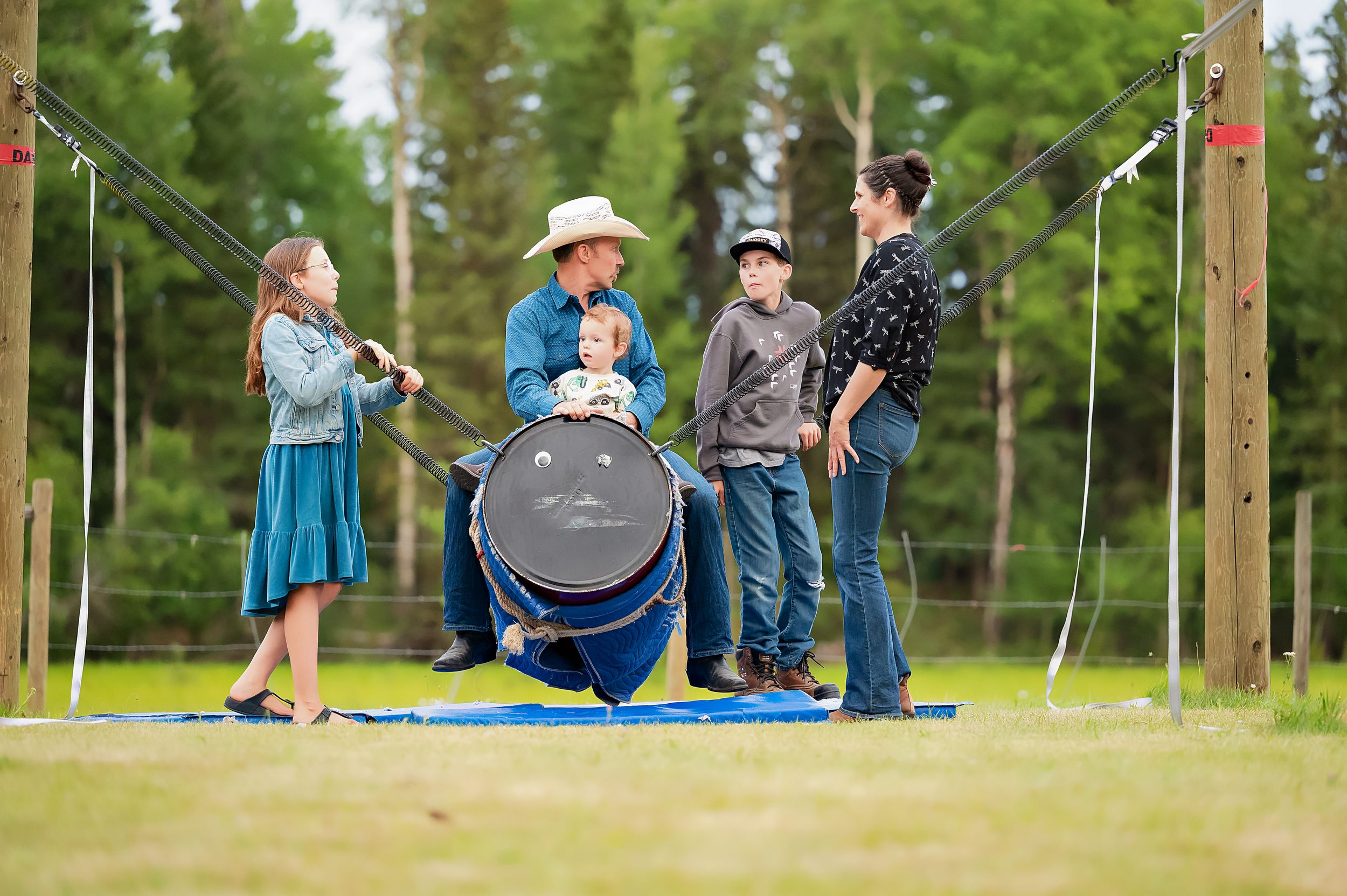 Group of people gathered around a grill in an outdoor setting with pine trees in the background.