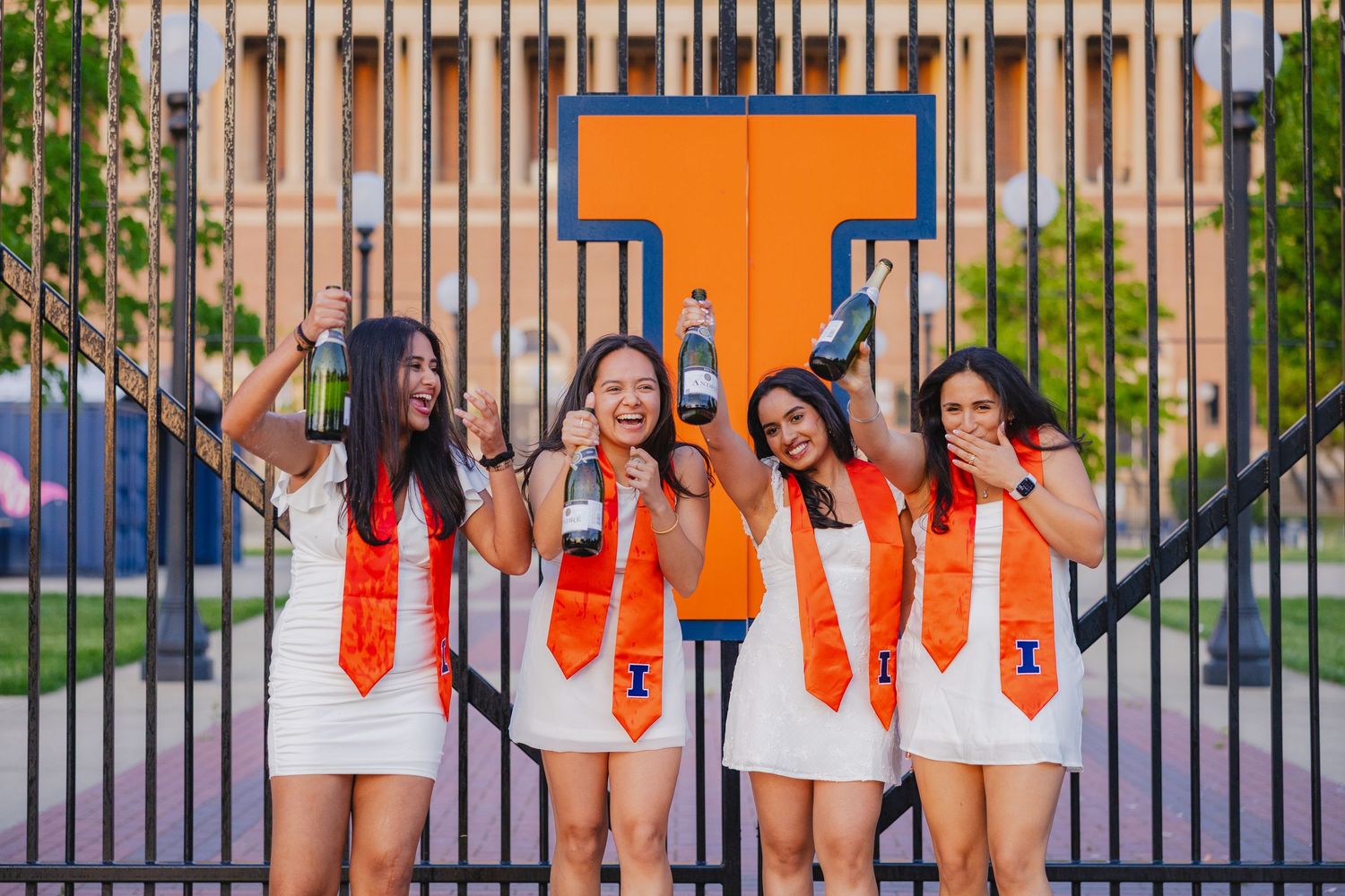 Four UIUC students with graduation stoles and champagne in front of Grange Grove gate
