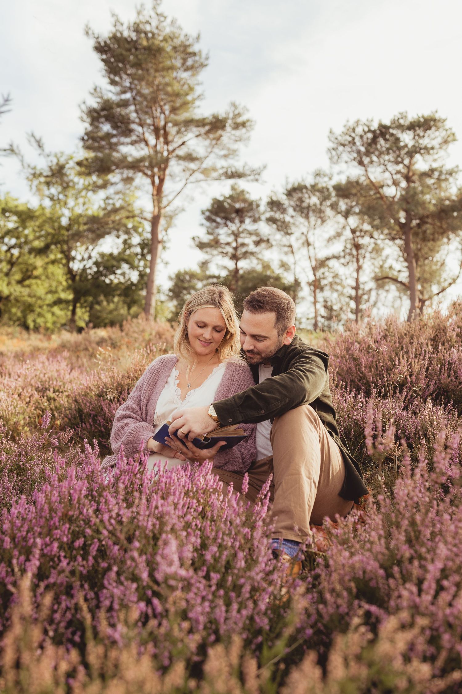 A couple holds ultrasound photos while sitting outdoors among pink flowering bushes in spring.