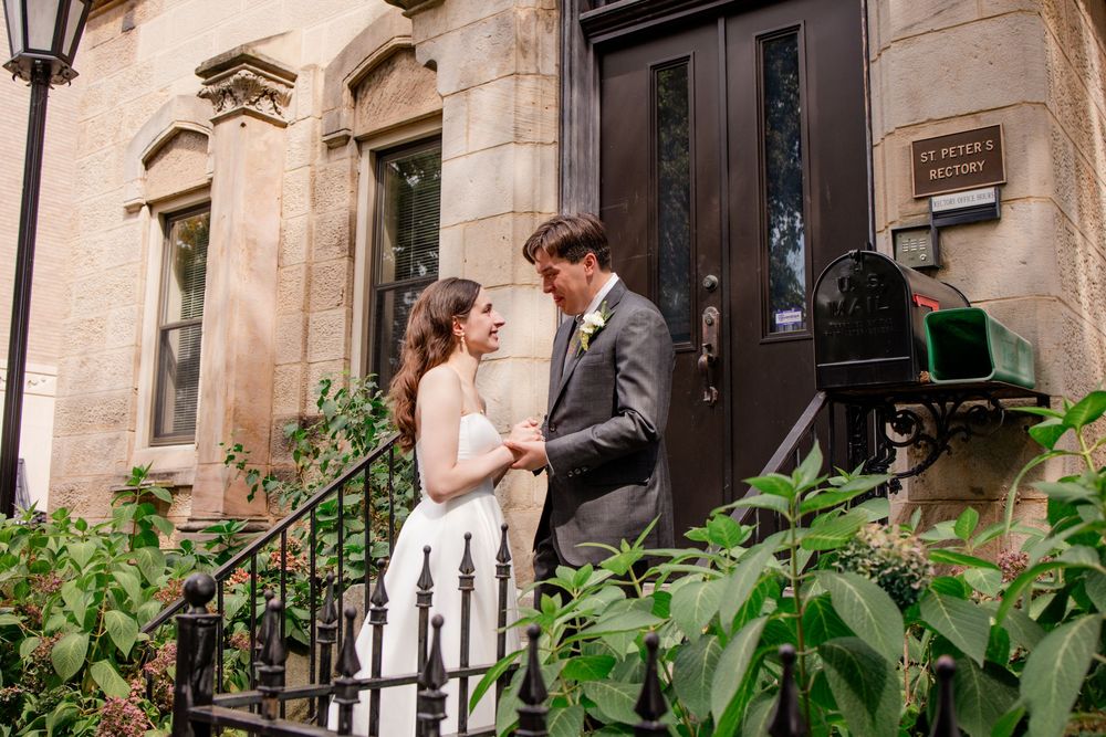A couple stands close together on the steps of a brownstone building with lush greenery surrounding the entrance.