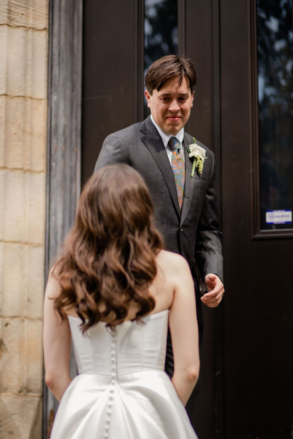 A bride in a white dress with button details on the back stands in front of a stone building entrance.
