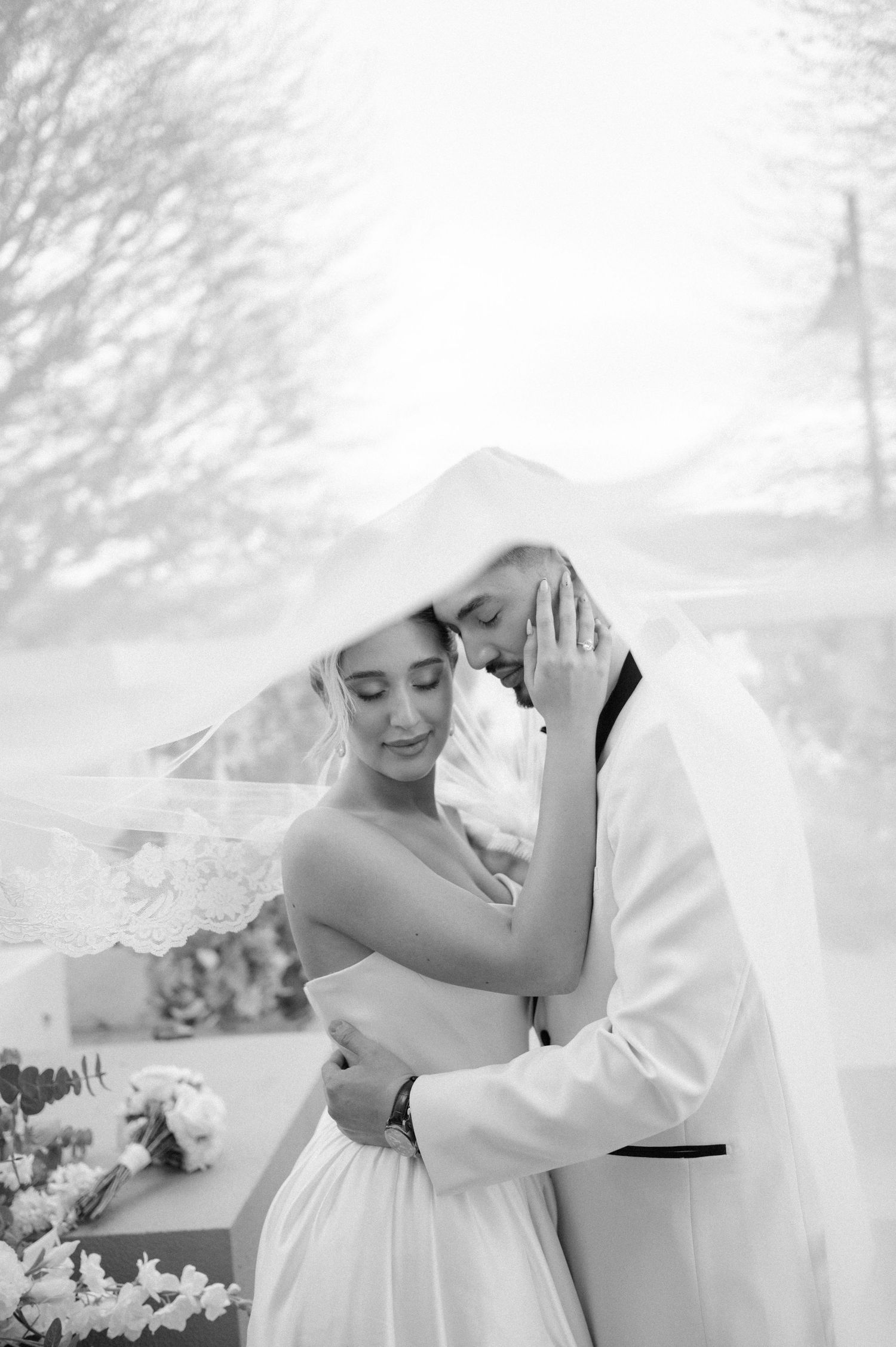 Black and white photo of bride and groom embracing tenderly under a flowing veil on their wedding day.