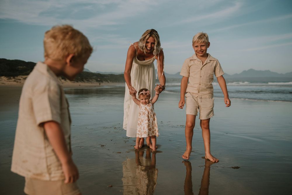 Candid beach scene of a mom guiding her little daughter’s first steps as her two sons laugh and play, reflecting joy, growth