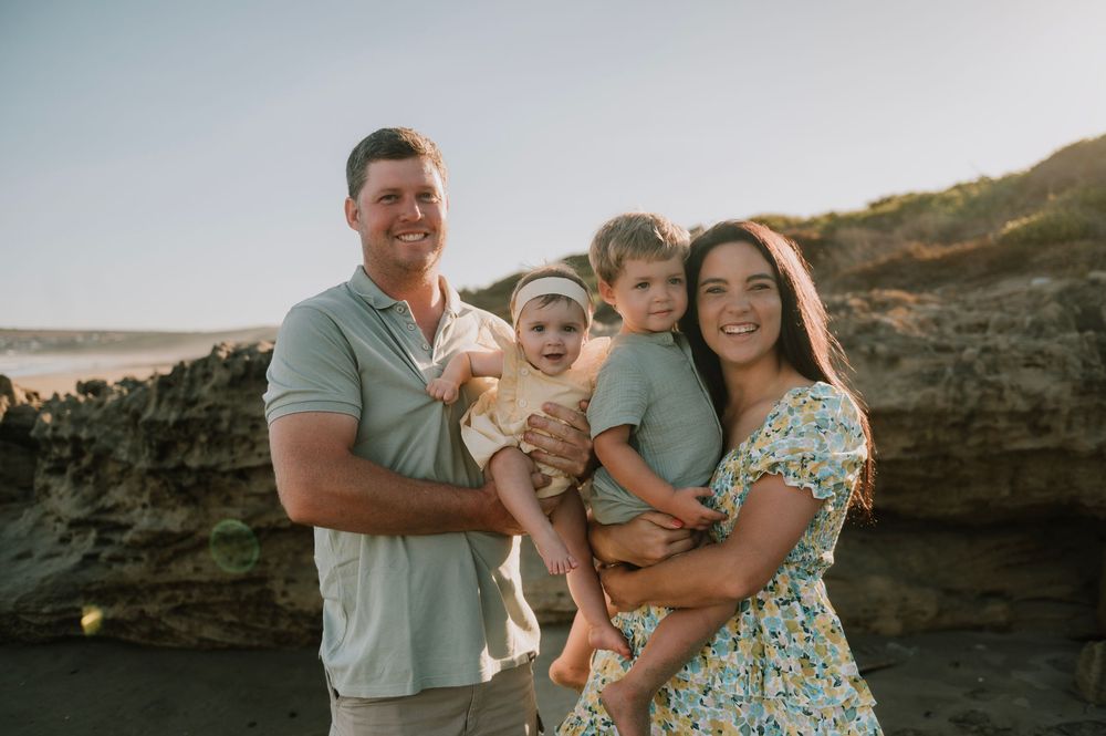 Candid golden hour family portrait at Boggomsbaai with parents and their two young children, set against rocky coastal formations