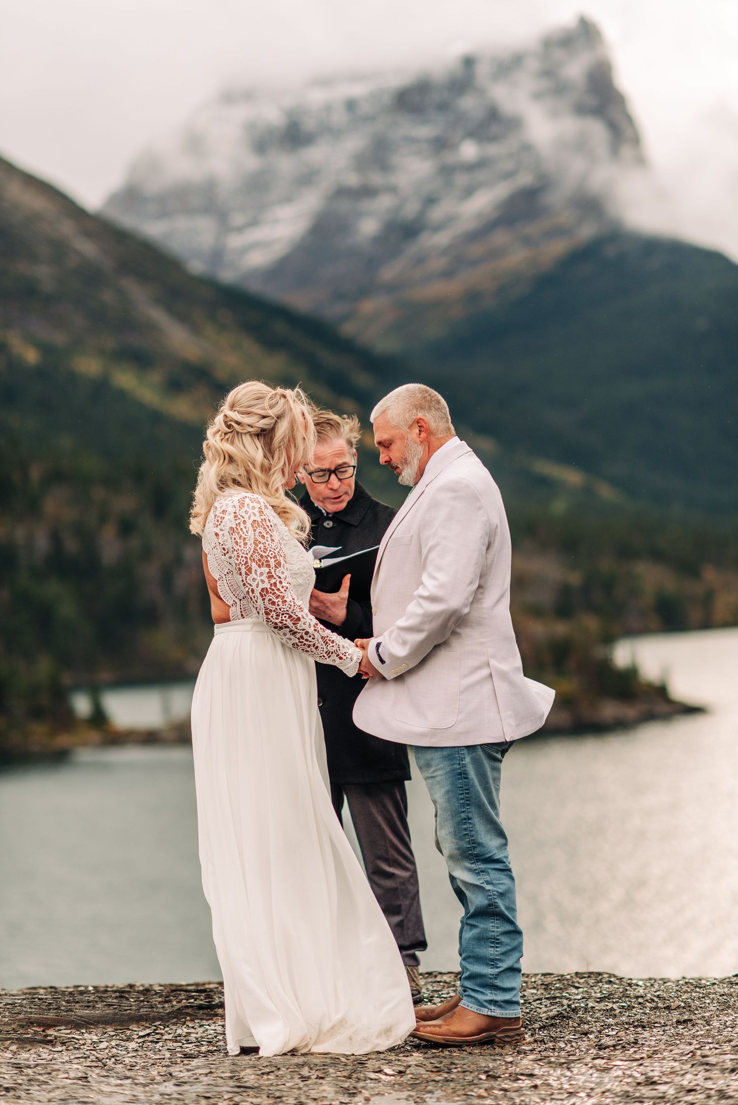 A couple shares an intimate wedding ceremony moment on a rocky beach with majestic mountain peaks in the background.
