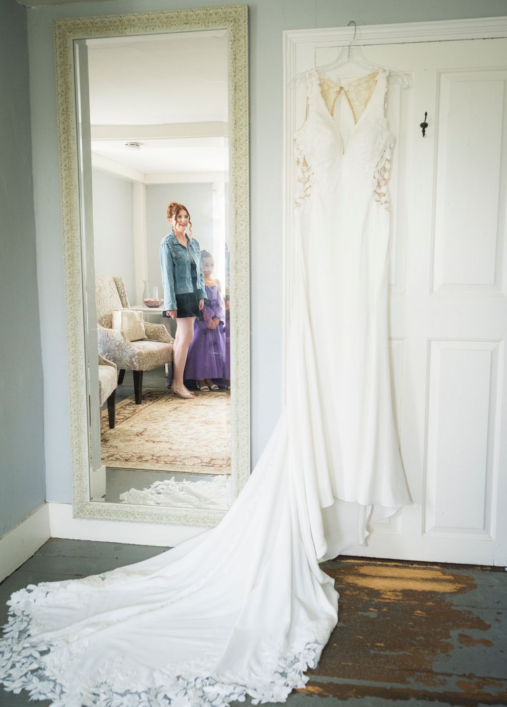 Elegant white wedding gown with lace train hangs on a door beside a full-length mirror.
