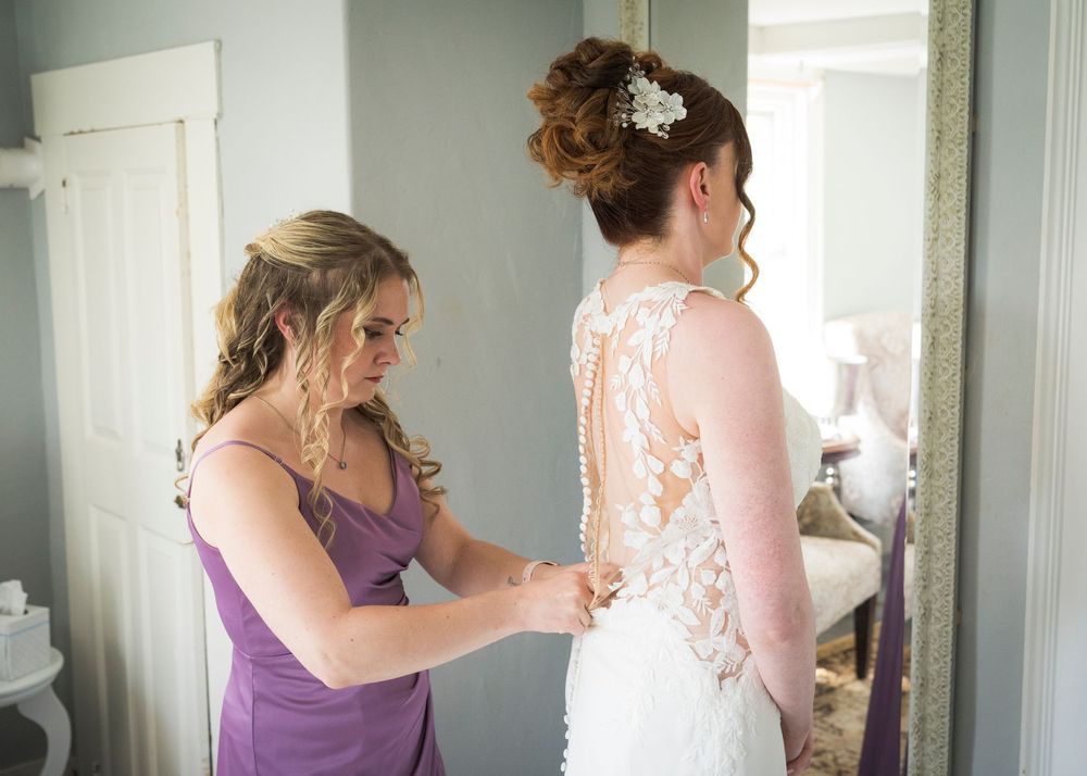 Bridesmaid in purple dress fastens buttons on brides lace wedding gown before ceremony.