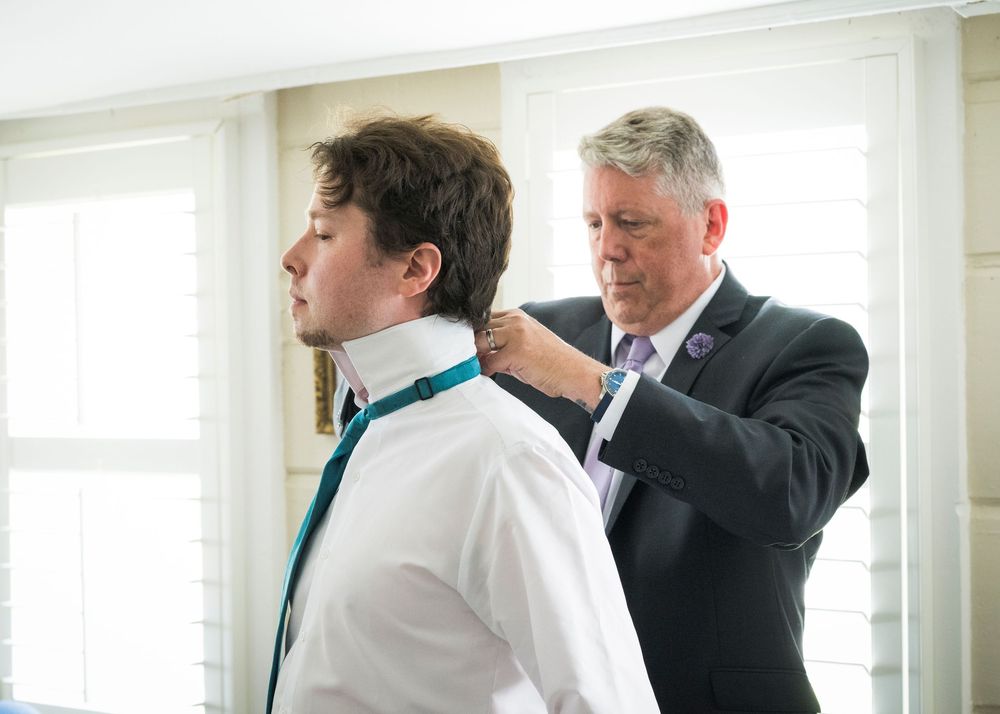 Father helps groom adjust teal tie before wedding ceremony in bright windowed room.