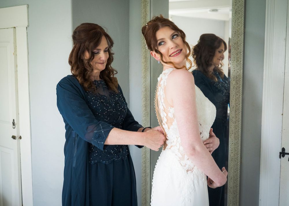Mother helps bride fasten lace wedding gown in front of mirror before ceremony.