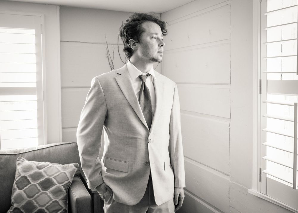 Groom in light gray suit gazing out window in black and white wedding portrait photo.