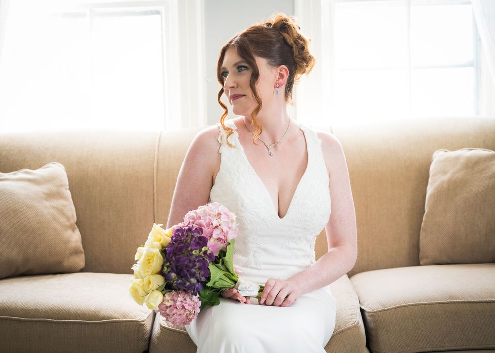 Bride in white gown holding colorful bouquet of pink, purple, and yellow flowers, seated on a beige sofa.