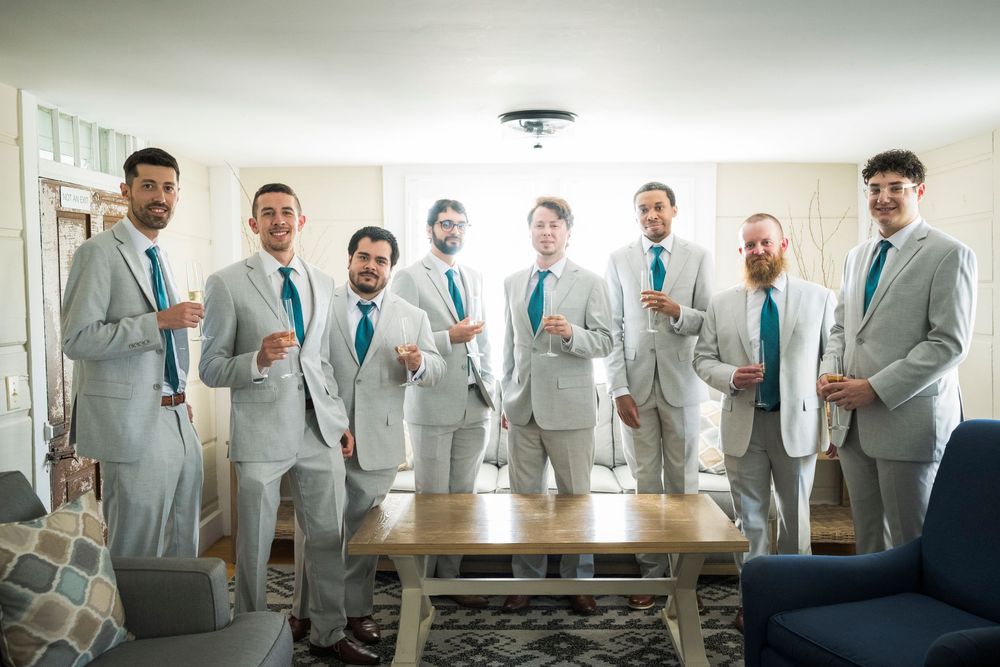 Groom and groomsmen in matching grey suits with teal ties pose together indoors before the wedding.