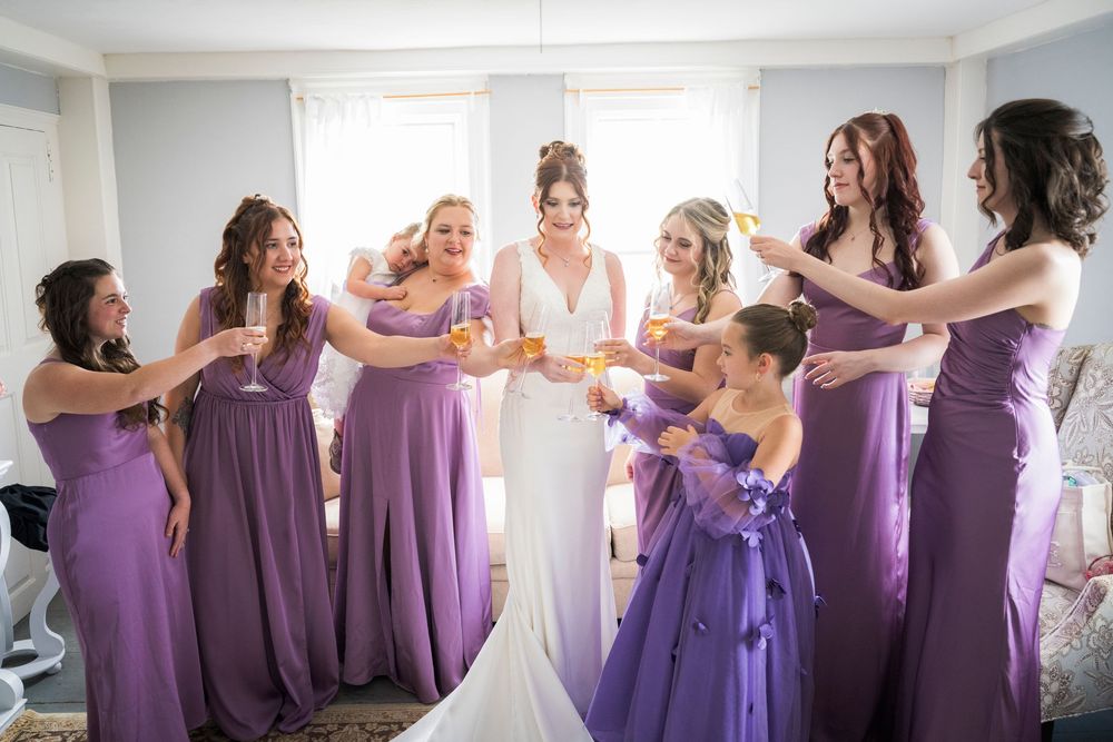 Bride in white gown toasts with seven bridesmaids in purple dresses before wedding.