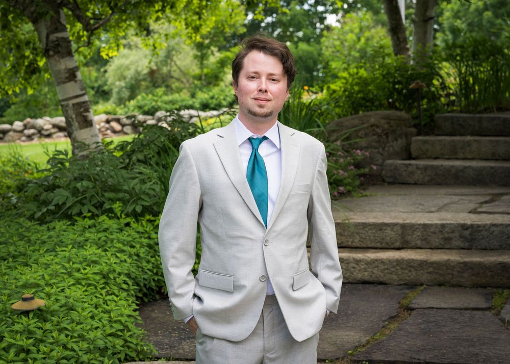 Groom in light gray suit with teal tie standing in a lush garden with stone steps and greenery.
