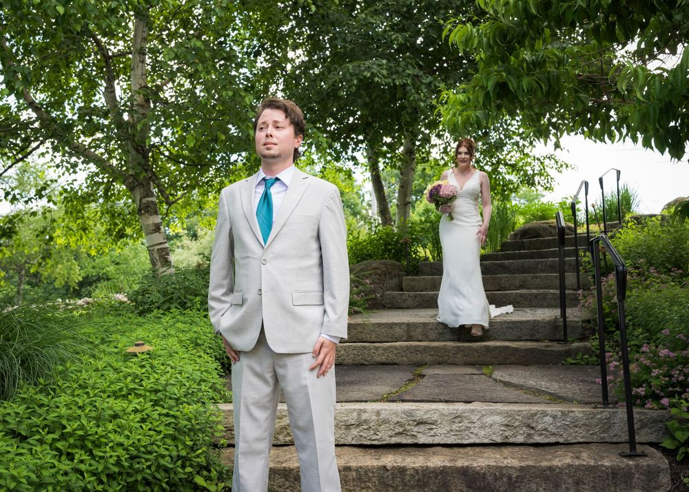 Groom in gray suit waits as bride descends stone steps in lush garden setting.