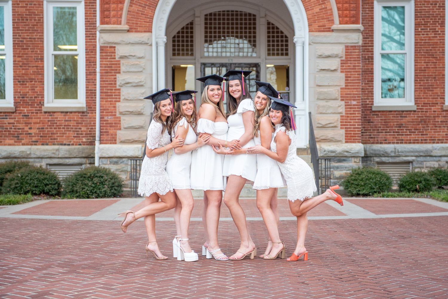 A group in matching white dresses and graduation caps pose playfully together on brick steps of a historic building.