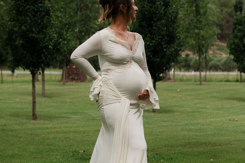 A pregnant woman in a flowing white dress poses gracefully in a lush green park setting.