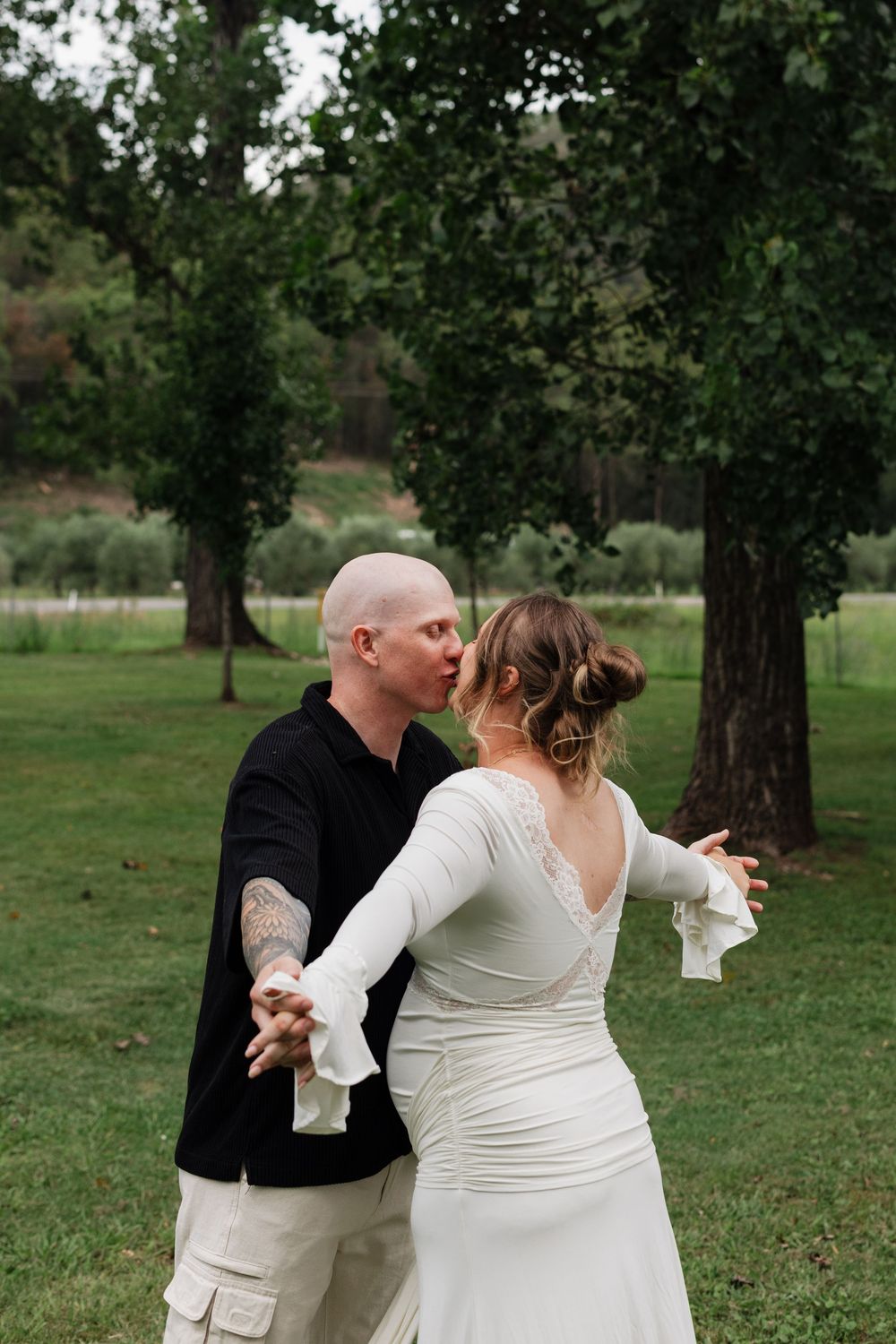 A couple in wedding attire dances together in a grassy park setting with trees in the background.