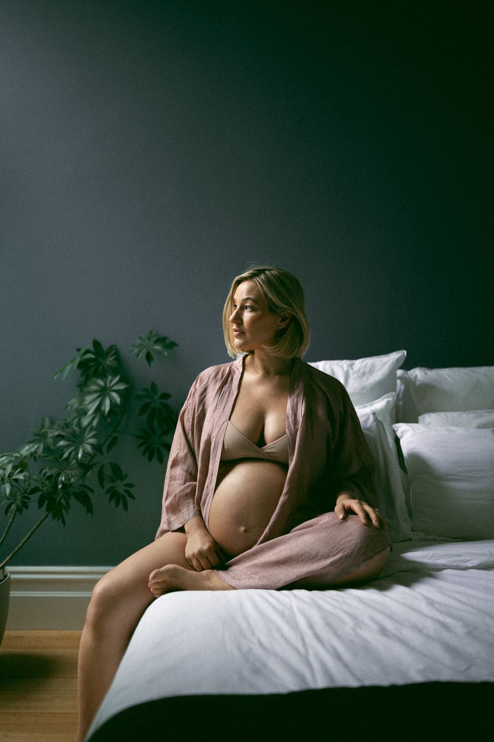 Artistic maternity photo series showing pregnant woman in soft lighting on white bedding with moody green backdrop.