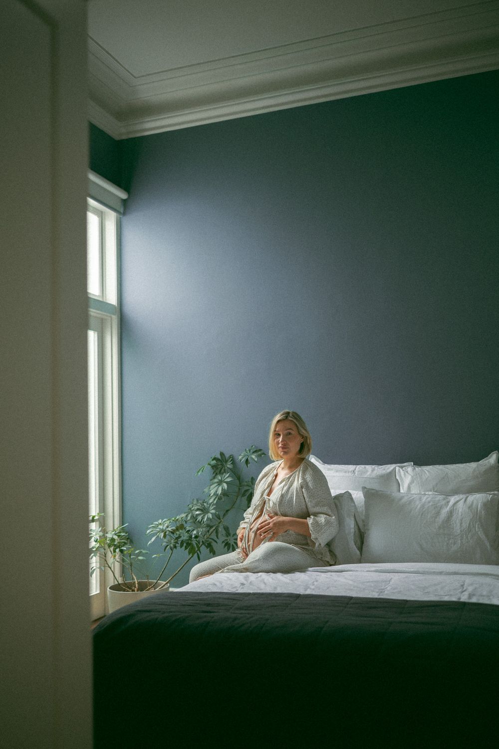 A person lies in a white bed against a dark teal wall with natural light streaming through windows.