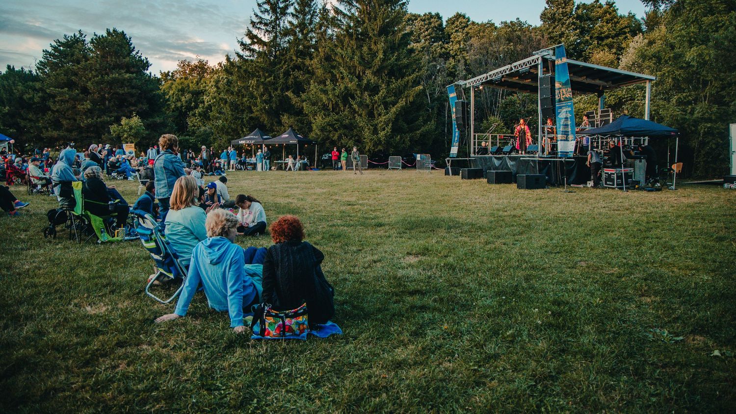 Outdoor concert audience sits on blankets in a grassy field watching performers on a small stage at sunset.