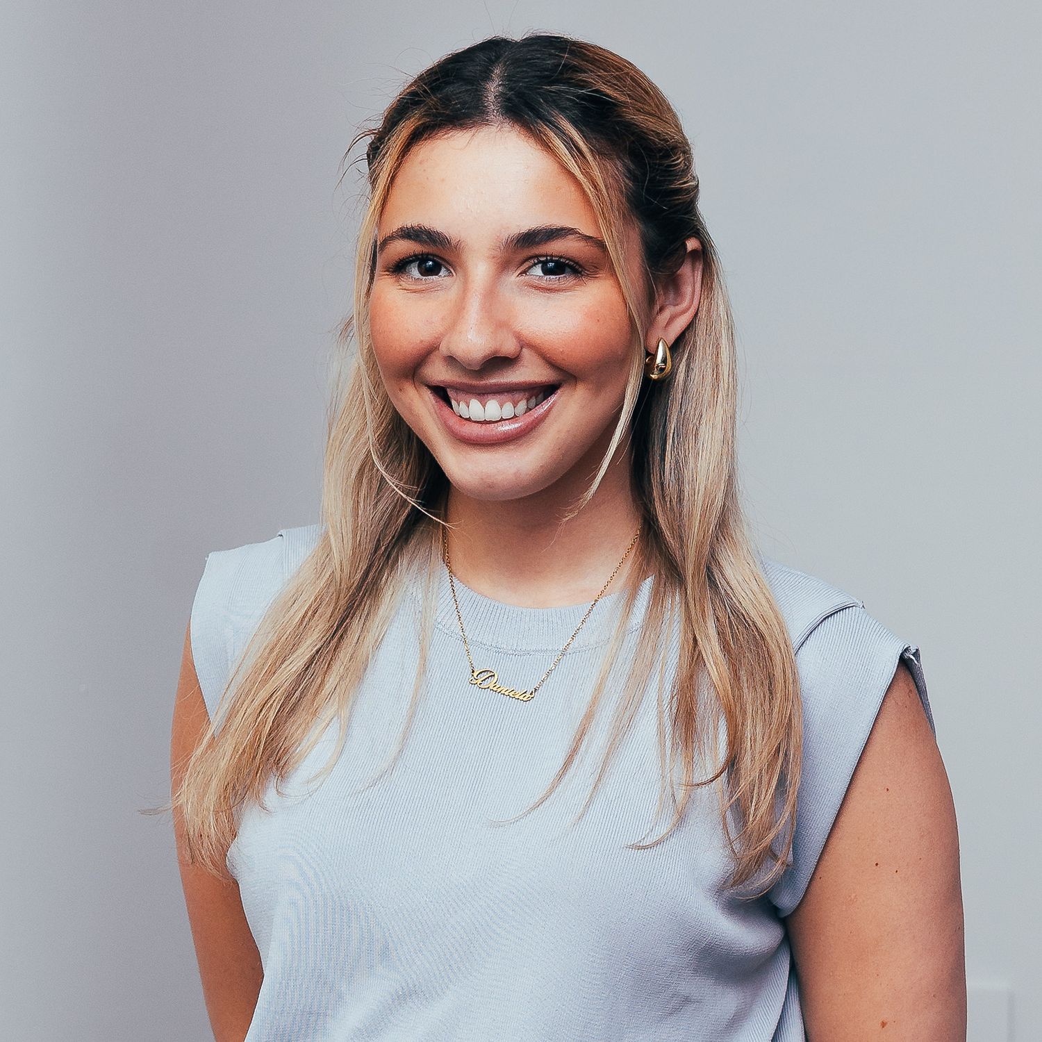 Professional headshot of someone with long blonde hair wearing a light gray sleeveless top and delicate necklace against neutral background.