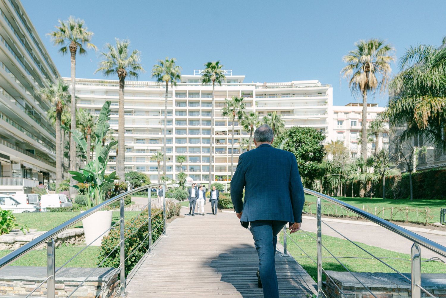 Person in blue jacket walking through a sunny beachfront property with palm trees and modern buildings in view.