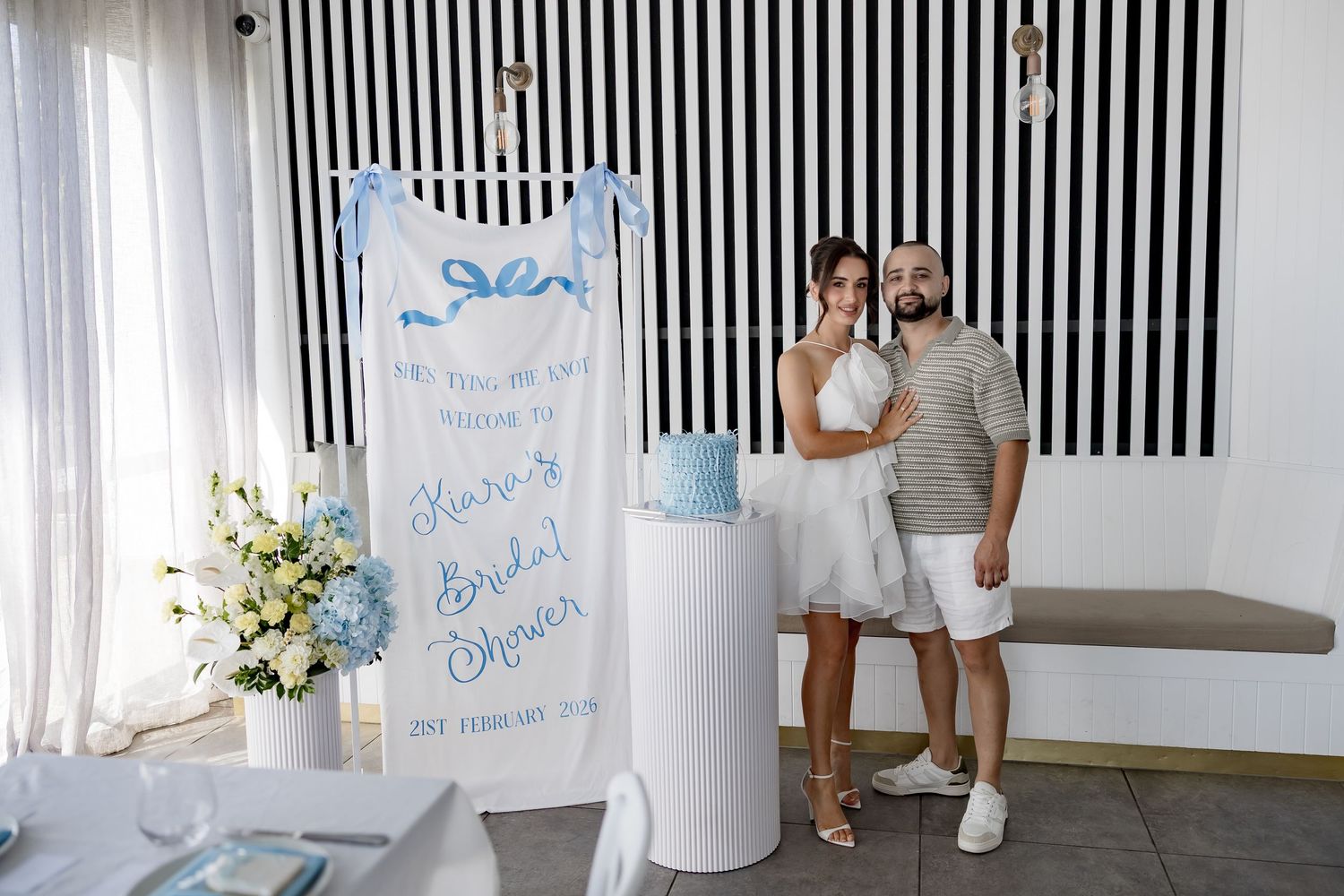Guests in white and light colored outfits gather at a bridal shower with black and white striped wall decor.