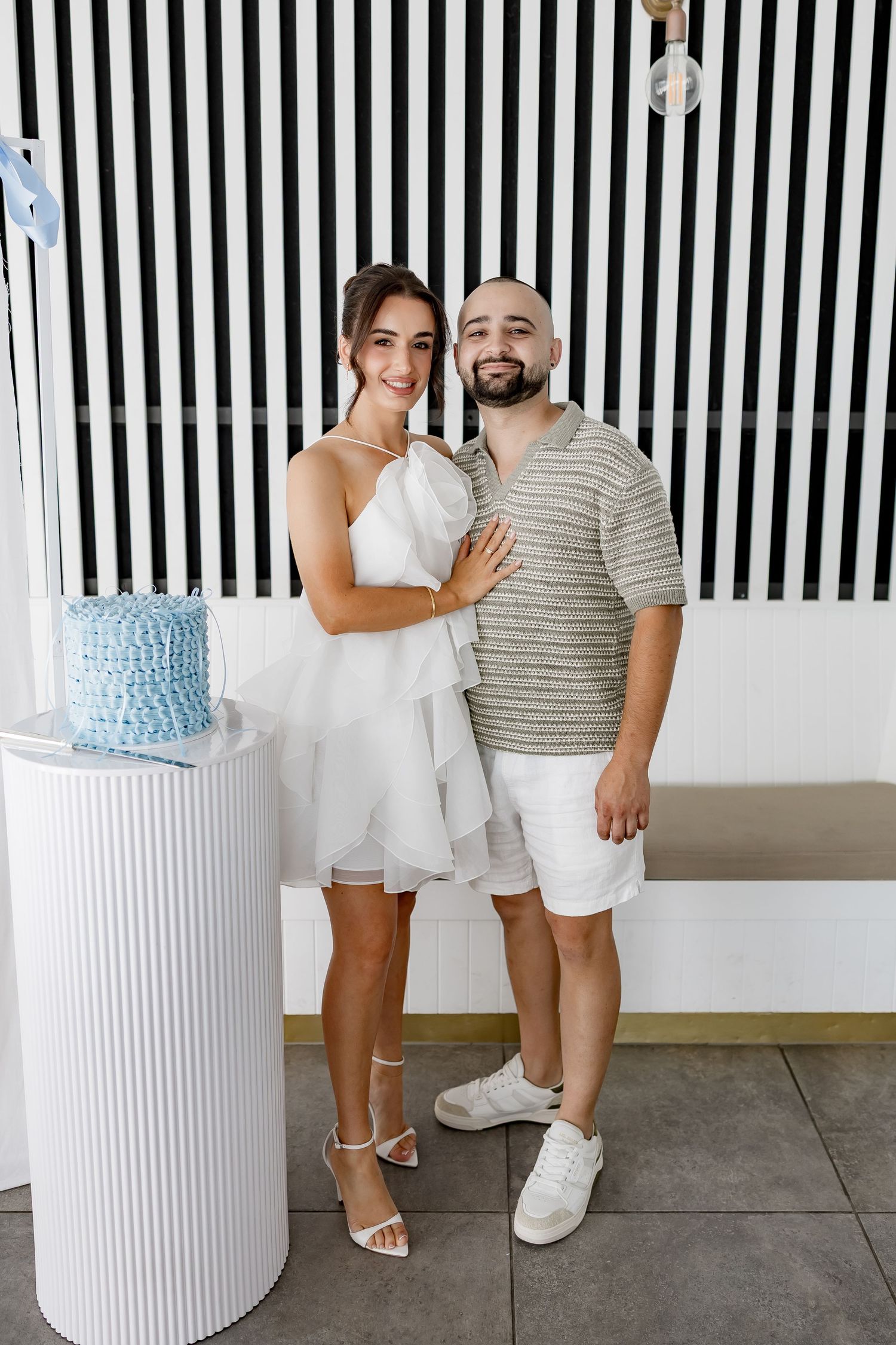 A couple stands next to a white pedestal with a blue and white tiered cake against a black and white striped wall.