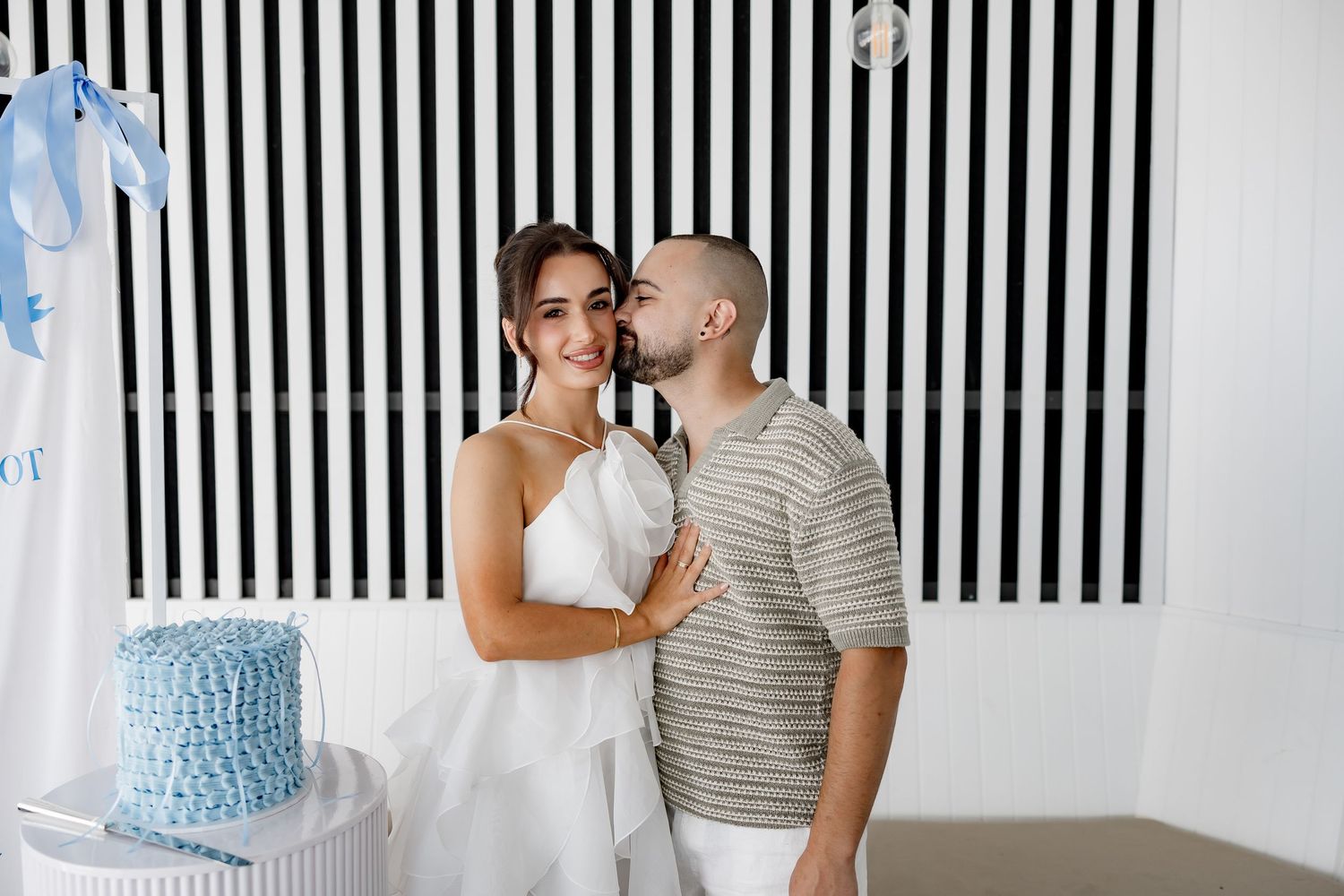 A couple poses together at a celebration with a black and white striped wall backdrop and blue cake display.