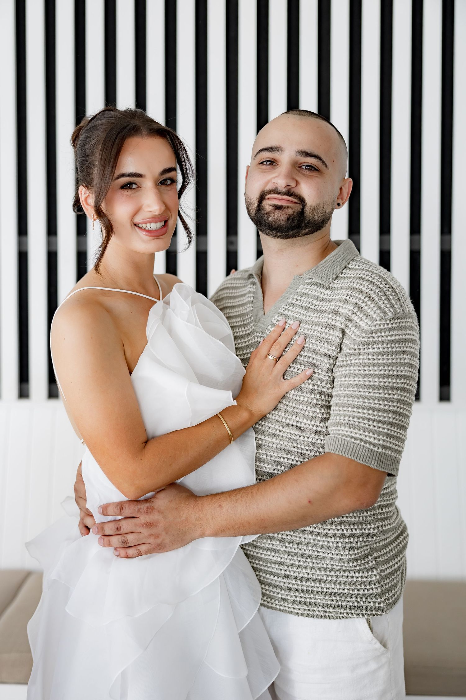 A couple embraces against a black and white striped wall, wearing white and patterned outfits.