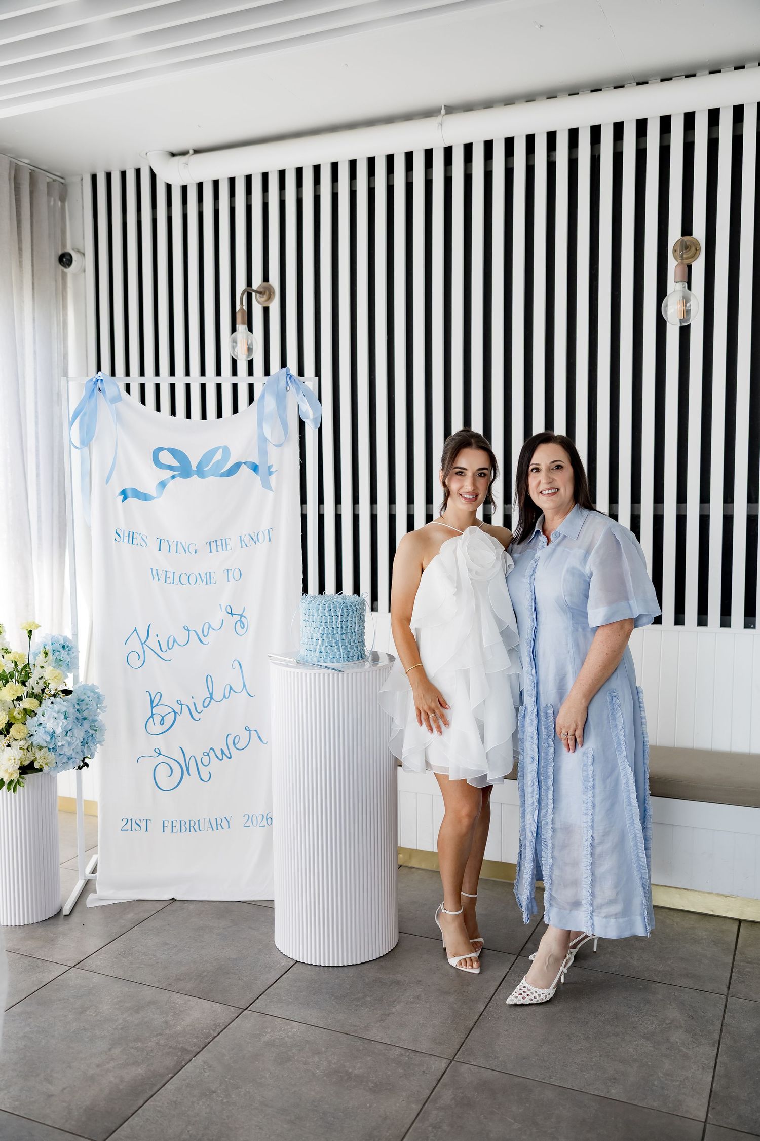 A bridal shower setup with blue decor, striped wall backdrop, and cake display on white pedestal.