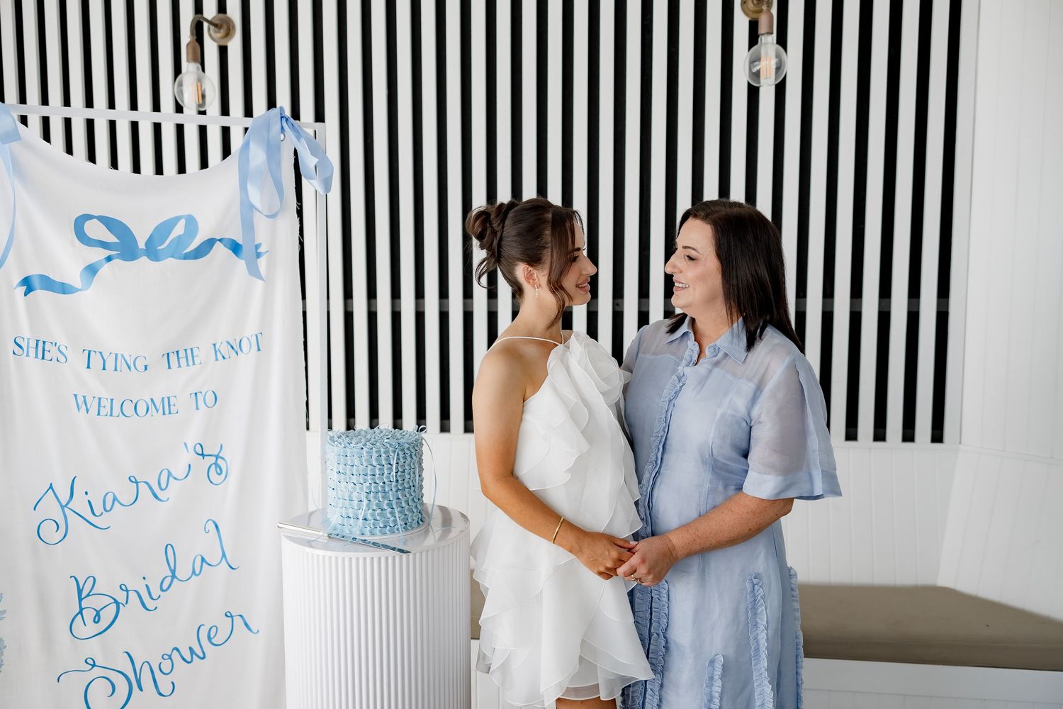 Two people stand together at a bridal shower event with blue and white decorations and striped backdrop.