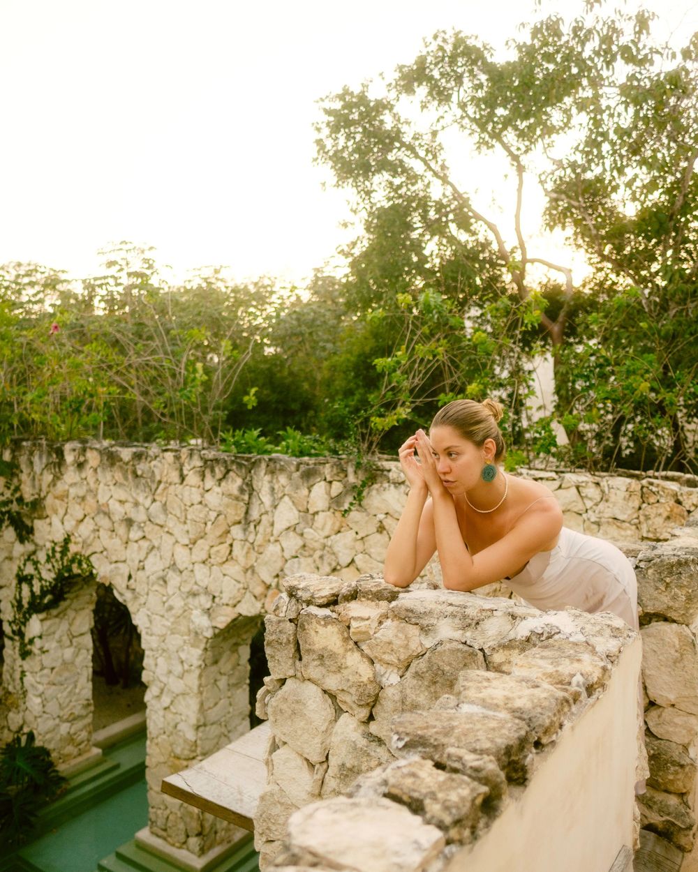 A person in a white gown sits thoughtfully on stone ruins surrounded by lush greenery in a dreamy outdoor setting.