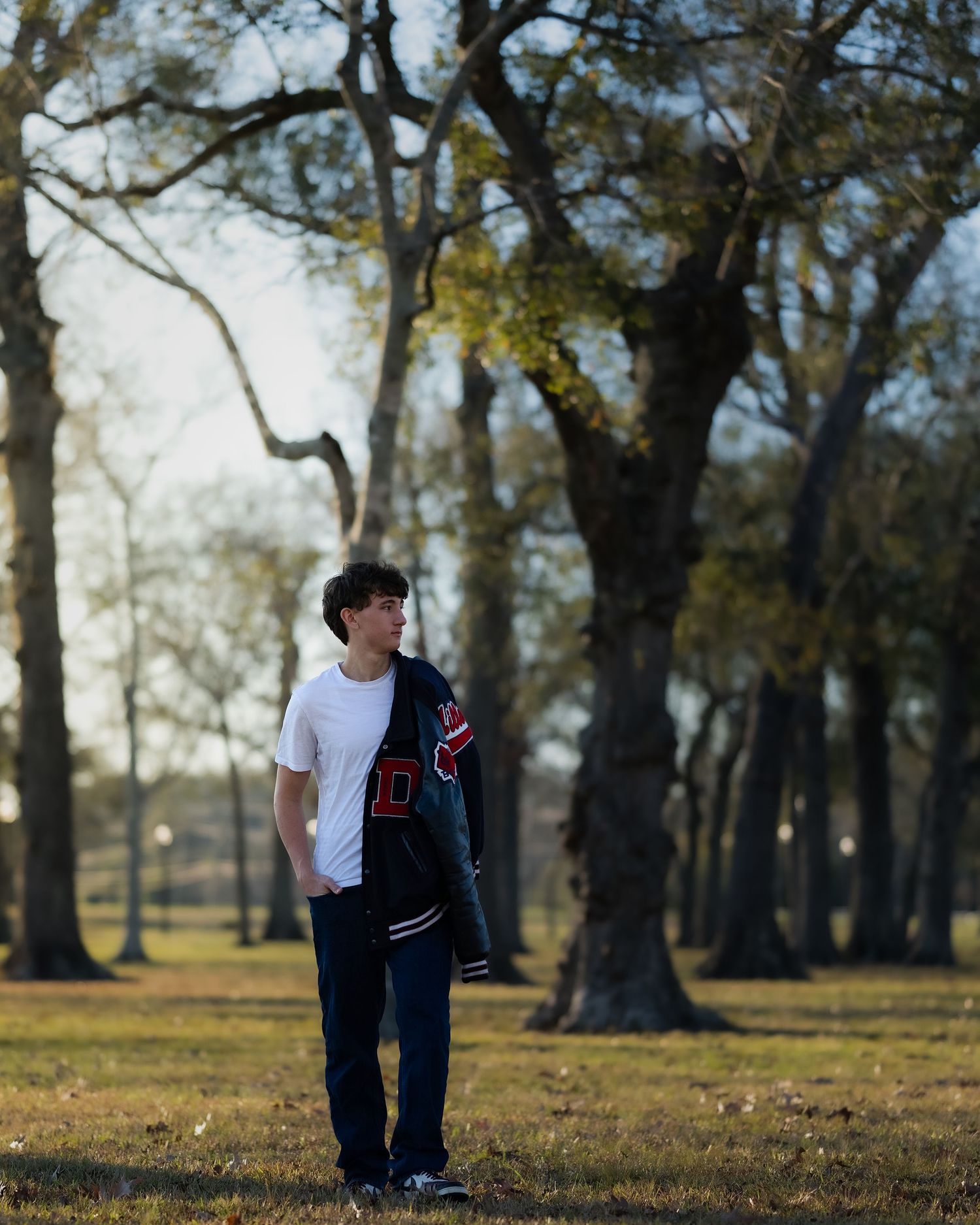A person in a varsity jacket stands in a wooded park area during golden hour lighting.