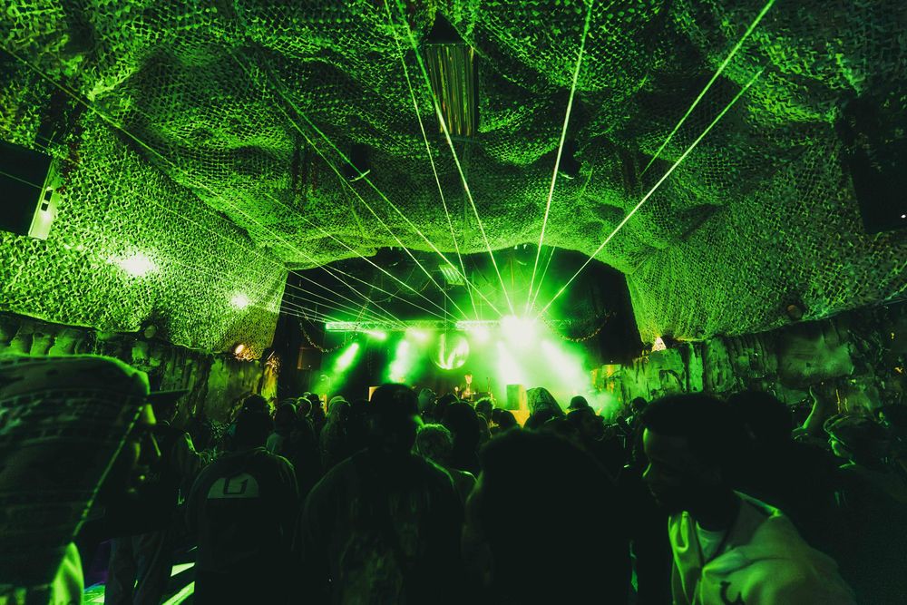 Green laser beams illuminate a dark underground cave tunnel with rocky walls and ceiling during a light show performance.