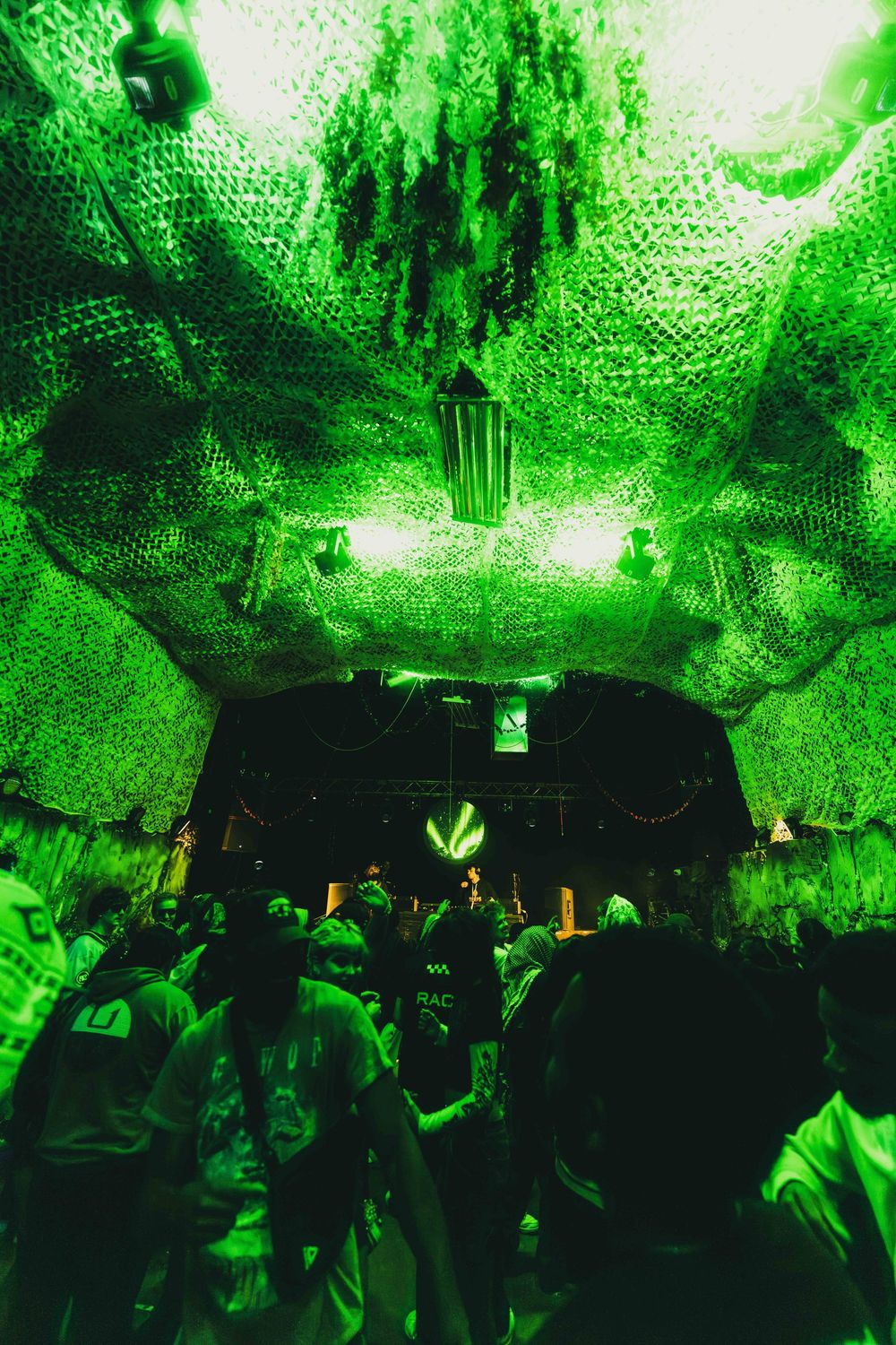 A nightclub crowd dances under vibrant green lighting and decorative mesh ceiling installations at a party venue.