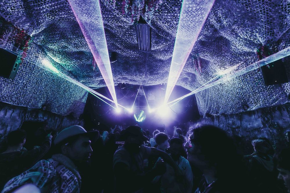 Purple and blue laser lights create a geometric pattern across a dark concert venue ceiling during a performance.