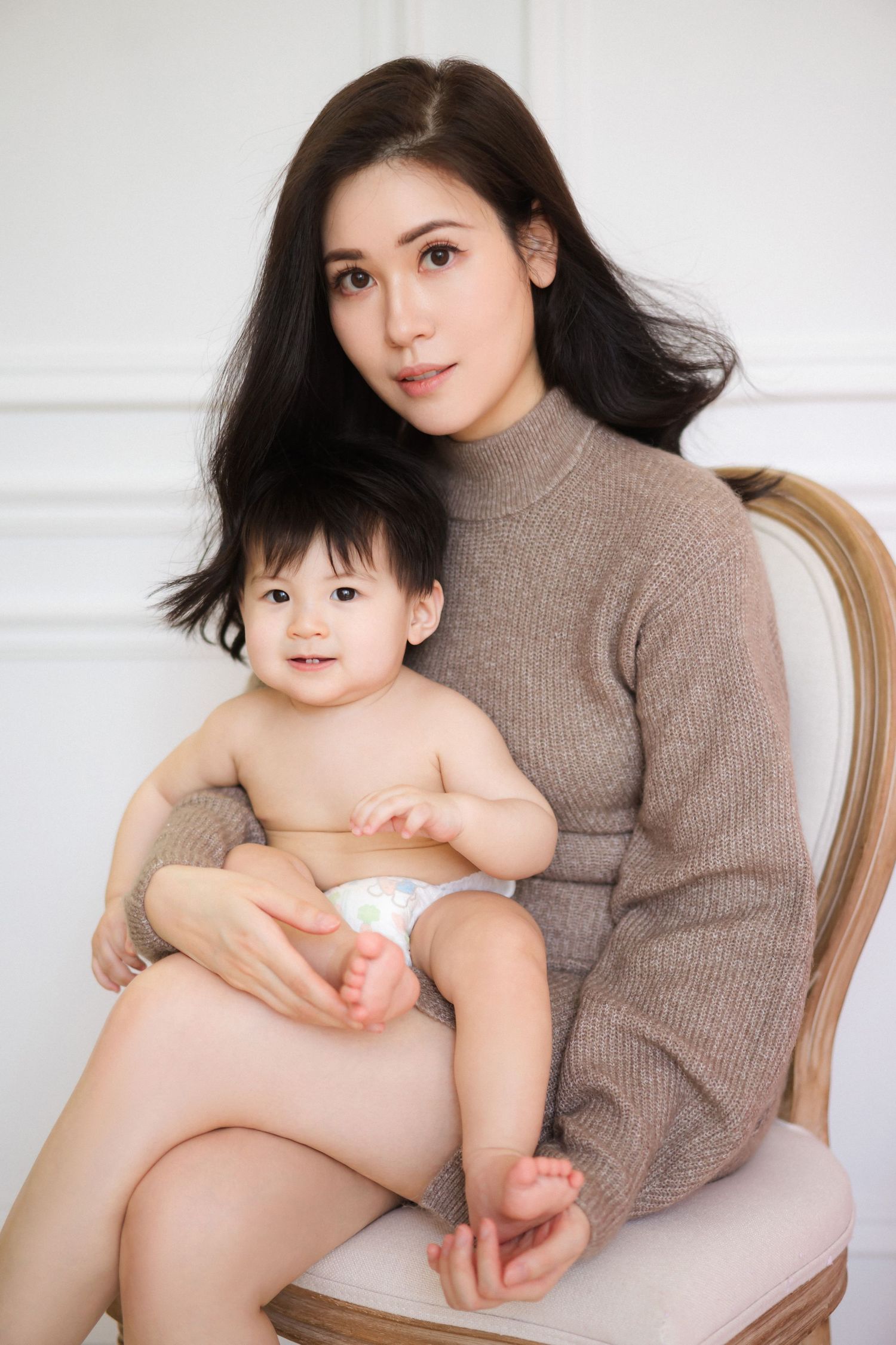 A parent and baby sit together on a beige chair, with the adult wearing a brown turtleneck sweater.一位家長和嬰兒並排坐在米色椅子上，大人穿著棕色高領毛衣。