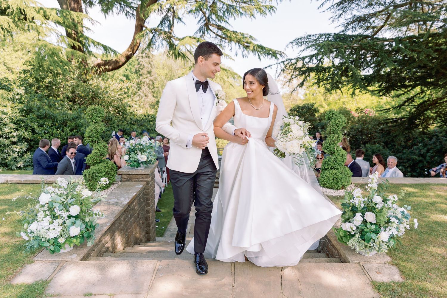 Couple in formal wedding attire walking through a lush garden setting during a summer outdoor celebration at Foxhills Manor