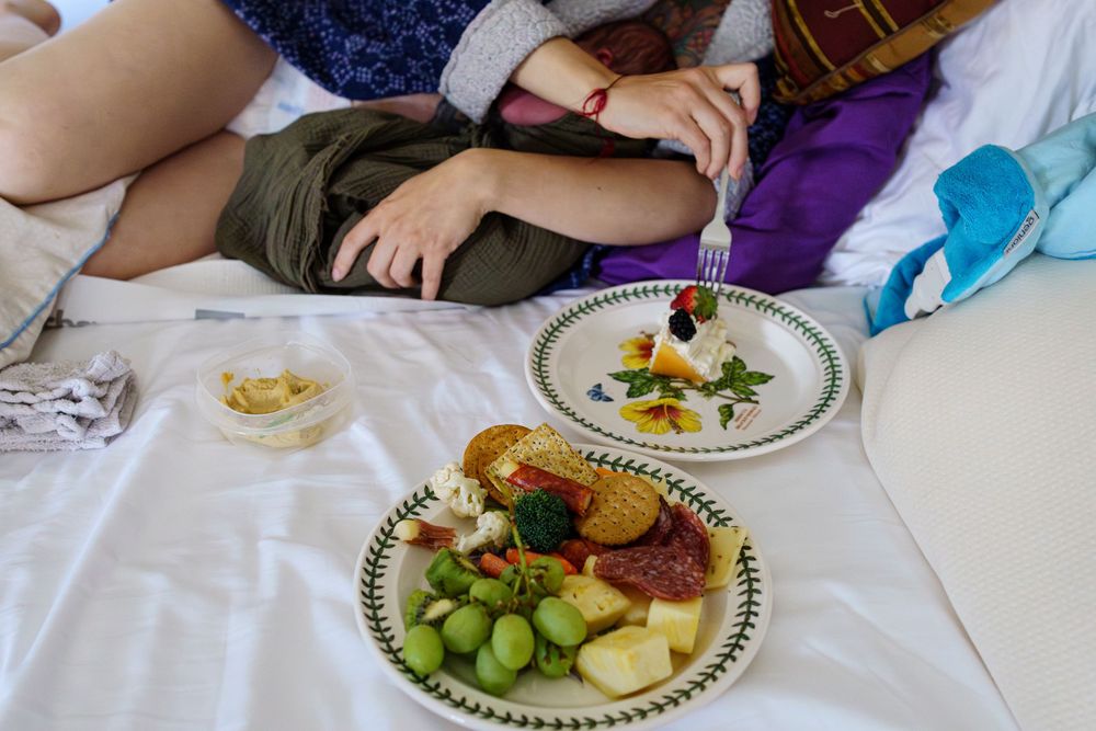 Two plates of food on white bedding show fresh fruit, salad, and sandwiches during a casual indoor meal.