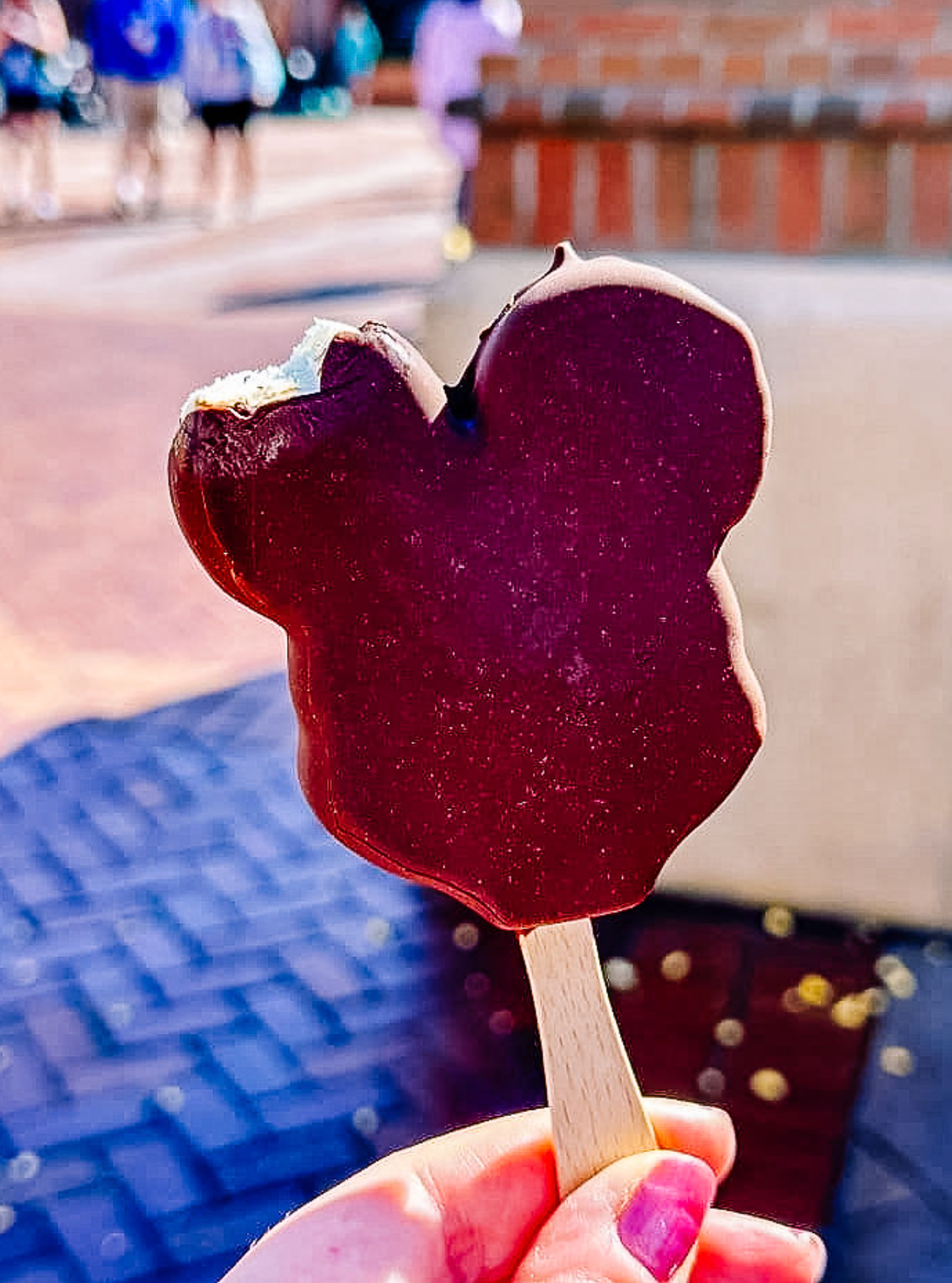 A chocolate-covered Mickey Mouse shaped popsicle ice cream treat held against a blurred outdoor background.