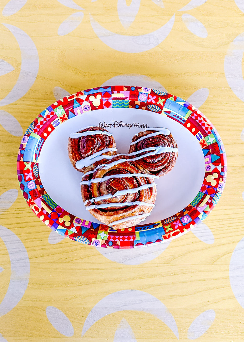 A cinnamon roll with white icing drizzle served on a Disney-themed paper plate with red and blue patterns.
