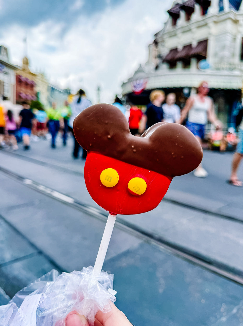 A Mickey Mouse-shaped chocolate covered ice cream bar on a stick with yellow button decorations at Disney theme park.