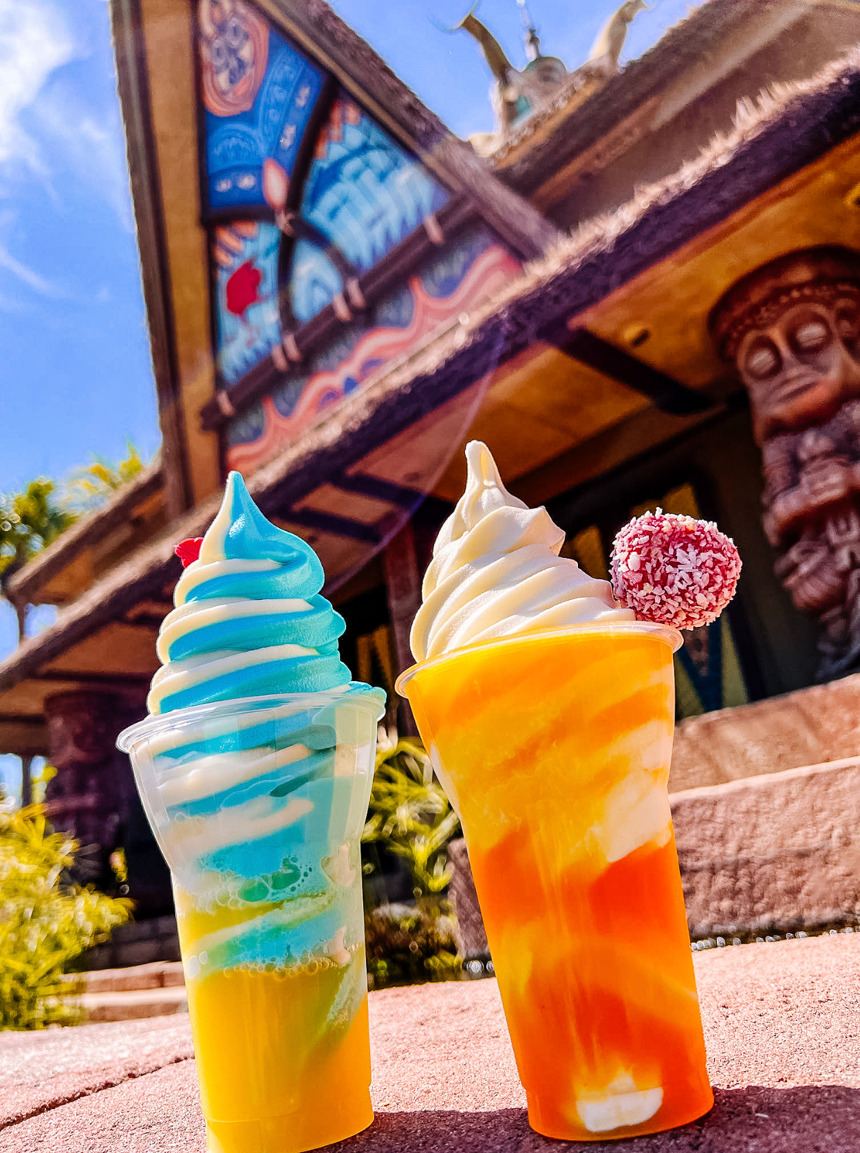 Two colorful frozen drinks with whipped cream toppings at a tropical themed tiki bar location in bright sunlight.